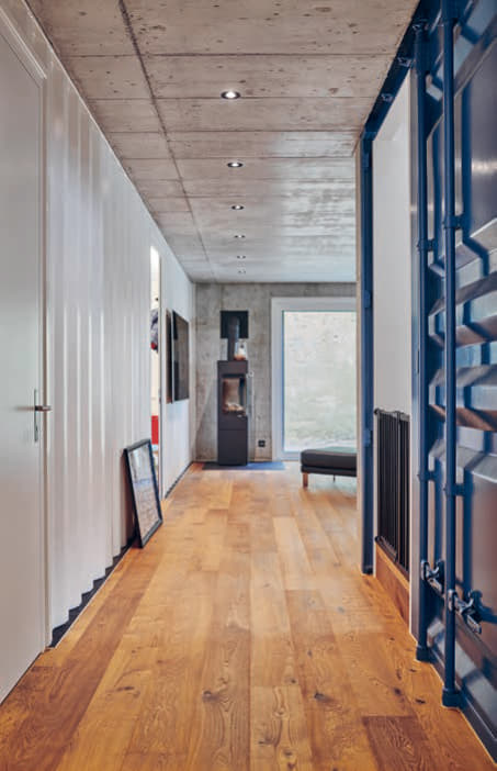 Ein warmer Holzboden führt zum Wohnbereich. Rechts rahmt die blaue Containertür die Treppe zum Obergeschoss. Hin- ter dem weissen Contai- ner links ist ein Kinder- zimmer und ein Reduit.