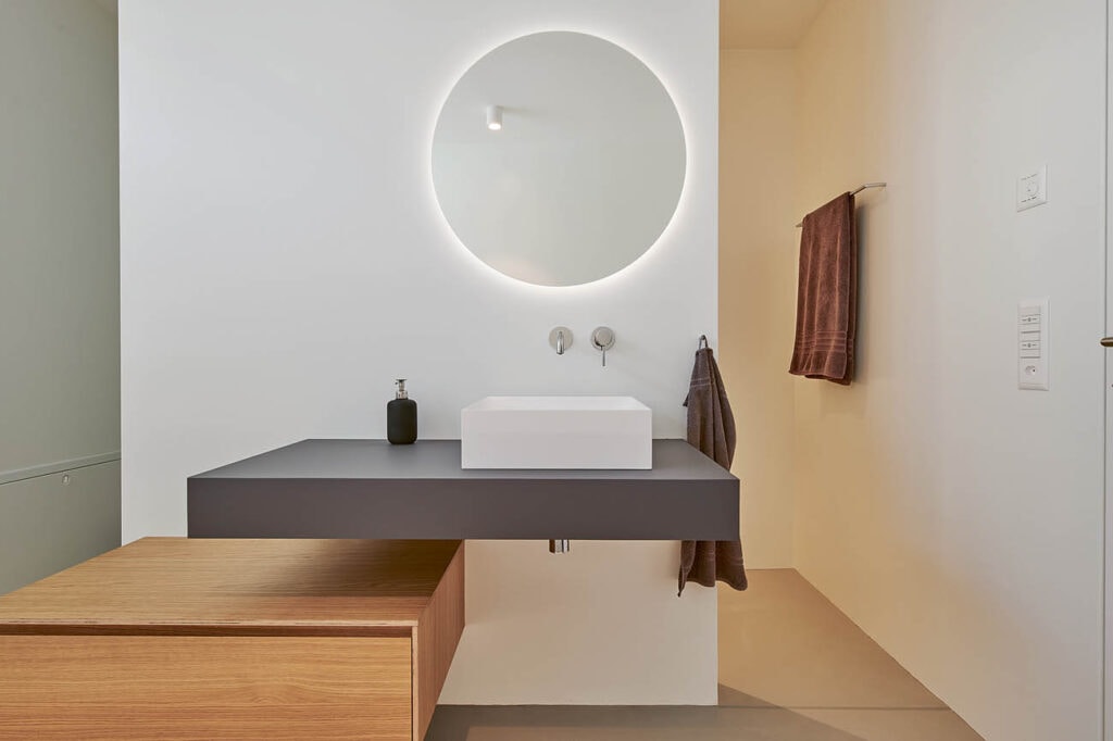 Badzimmer mit anthrazit Ablageflächen und einem runden Spiegel.